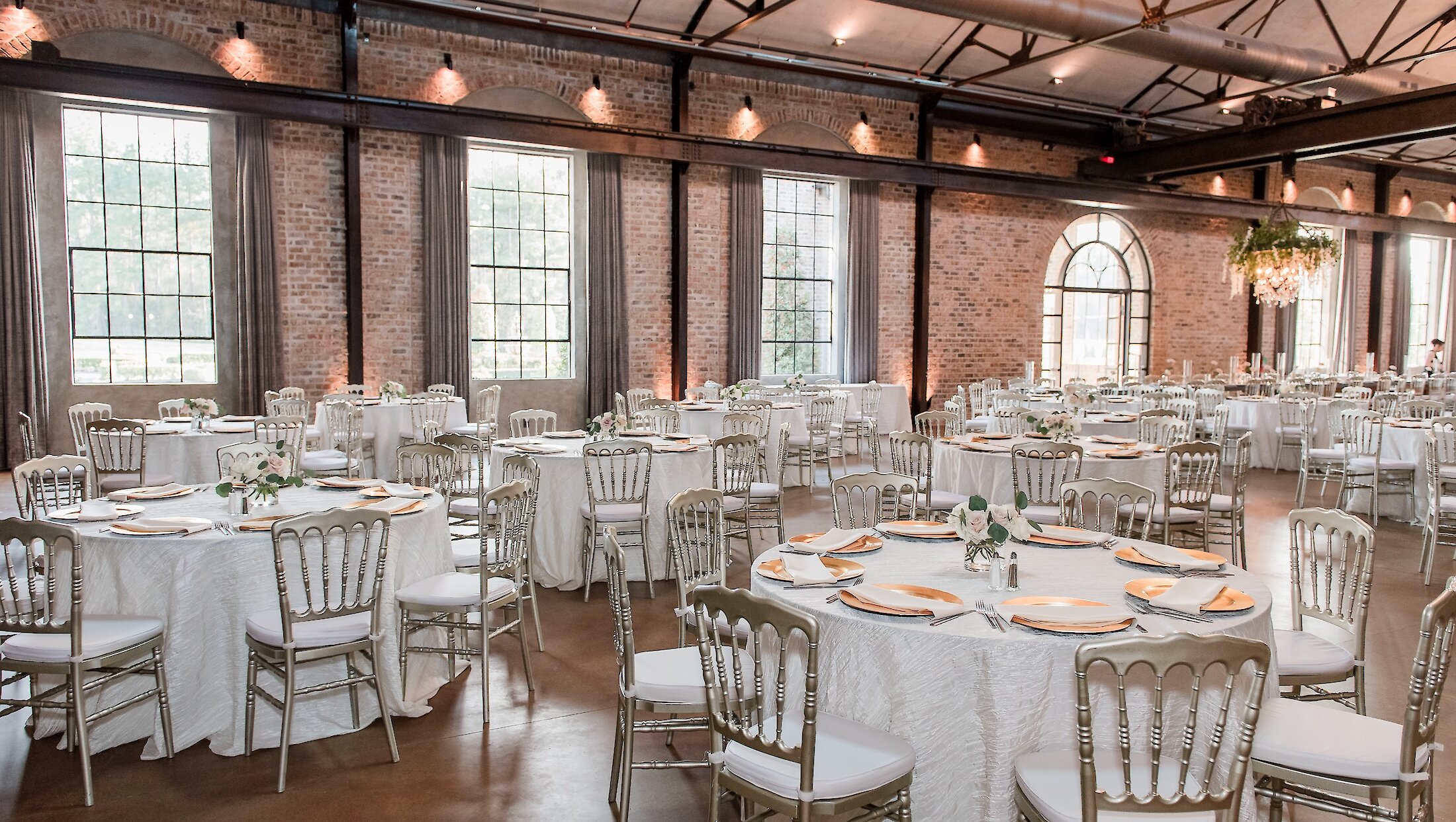 An expansive wedding reception hall featuring high ceilings, exposed beams, brick walls, and silver chiavari chairs at round tables.