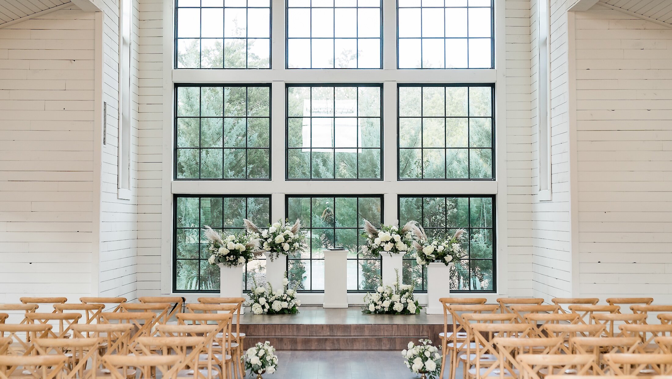 A bright, rustic indoor ceremony space with white shiplap walls, wooden cross-back chairs, and large windows.