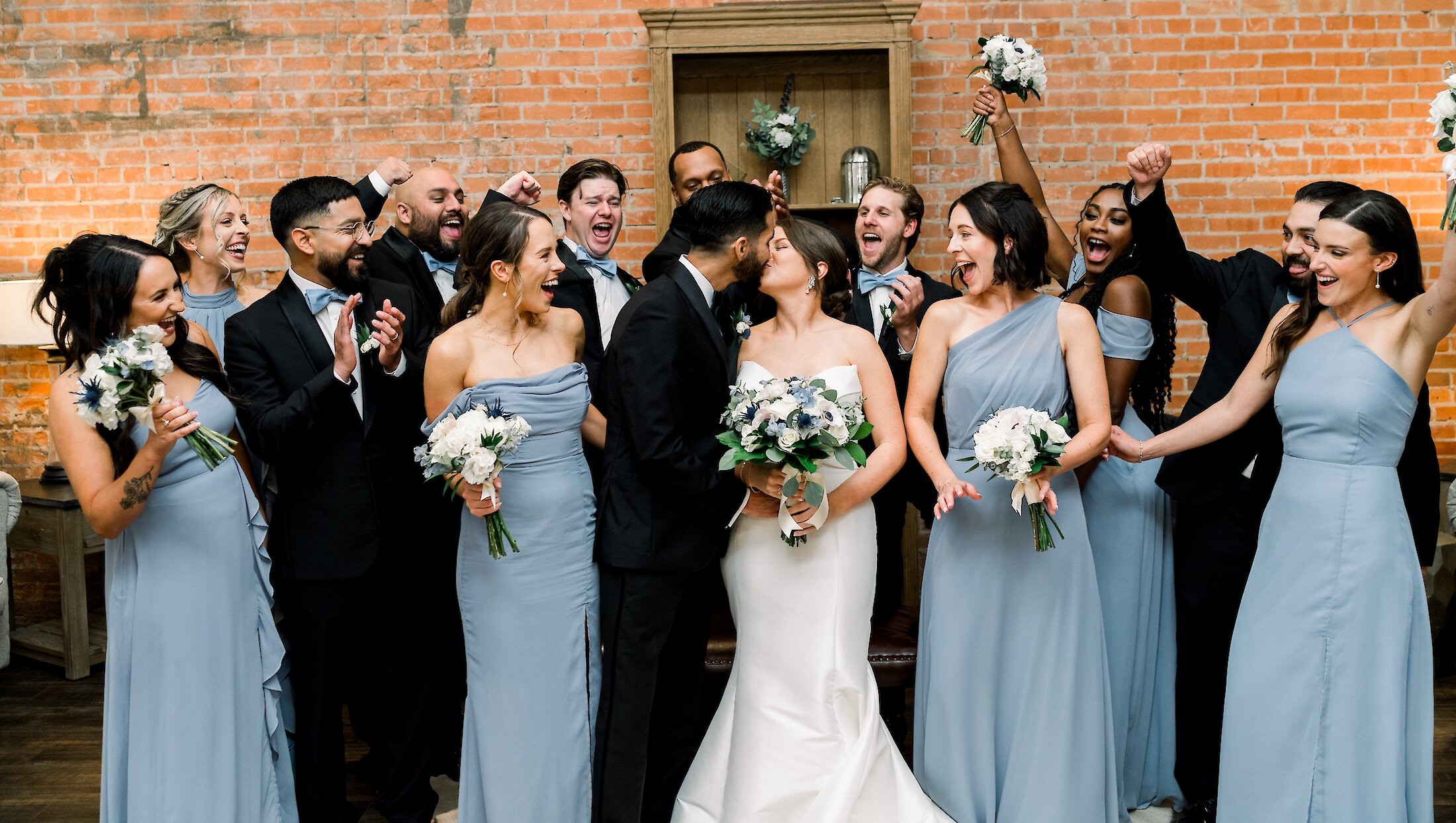 The ecstatic wedding party surrounding the bride and groom for a fun group photo against a brick wall.