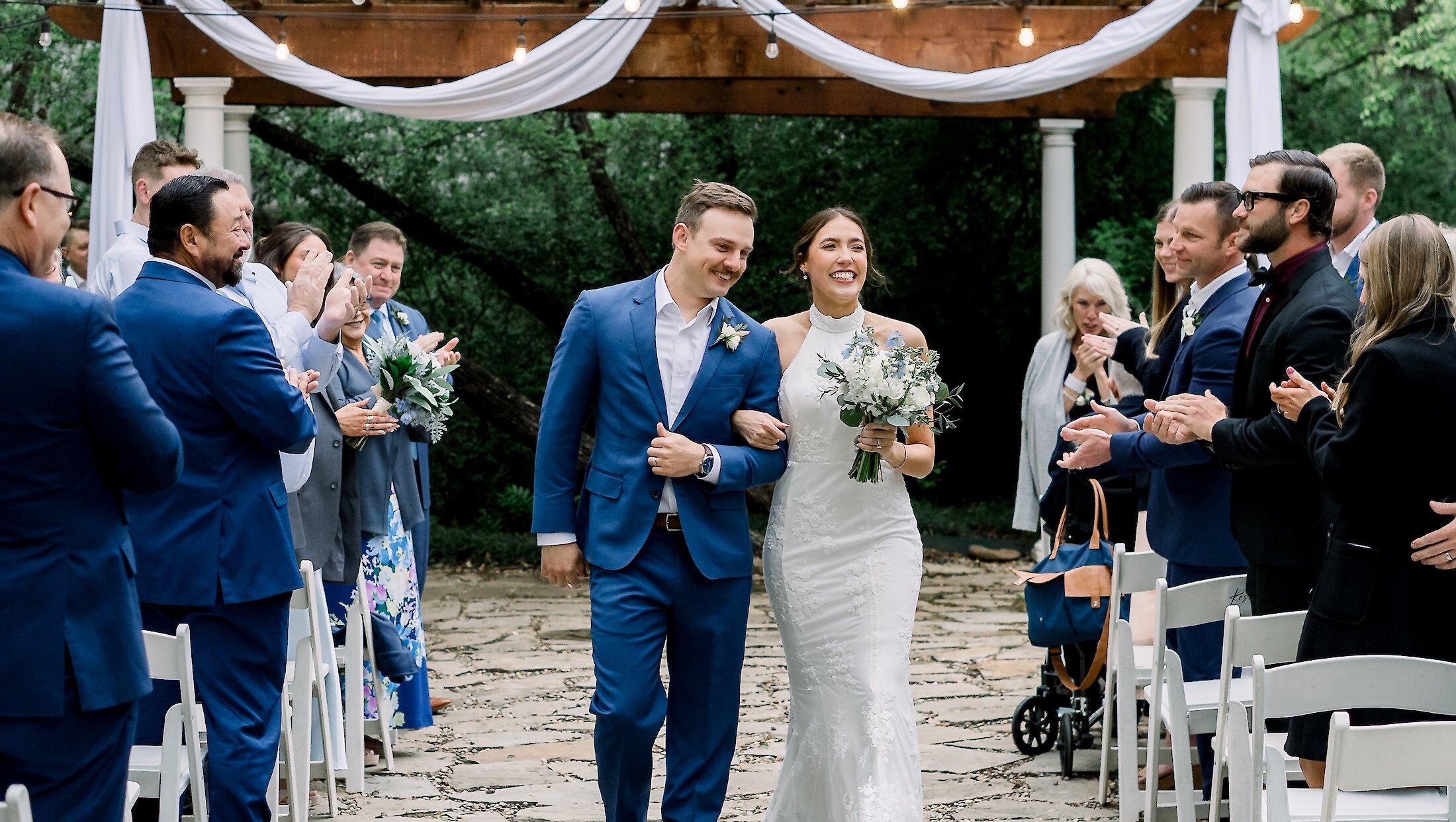 The bride and groom smile while walking arm-in-arm down the outdoor aisle as guests clap beneath a wooden pergola draped with white fabric.