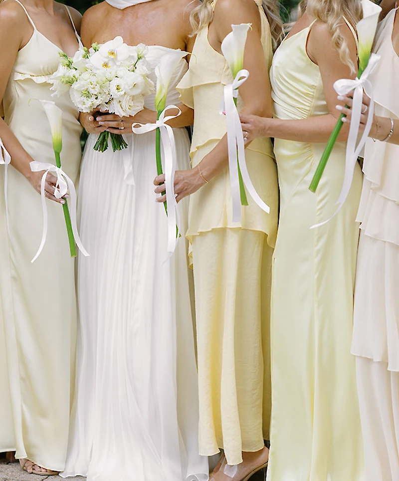 A wedding flat lay featuring the invitation suite, photo booth strips, rings, lemons, calla lilies, and green floral accents on a white surface.