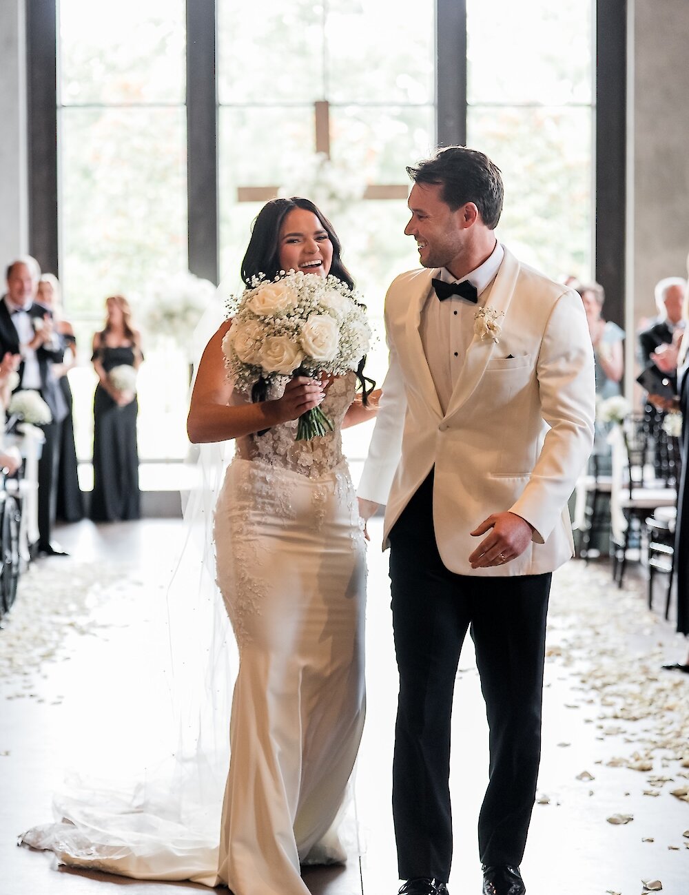 A laughing bride holding a white rose bouquet walks down the aisle with her groom, who is wearing a white tuxedo jacket and black pants, after the ceremony.