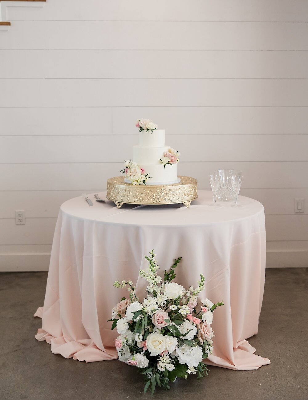 A simple three-tier white wedding cake decorated with pink flowers sitting on a gold stand atop a table with a blush pink tablecloth.