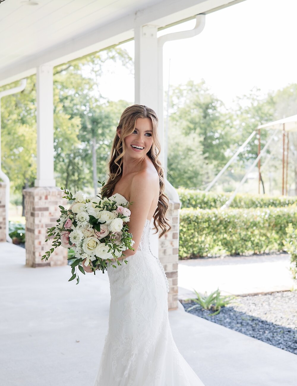 A beautiful smiling bride in a strapless lace wedding dress holds a large bouquet of white and blush pink flowers on a covered outdoor porch.