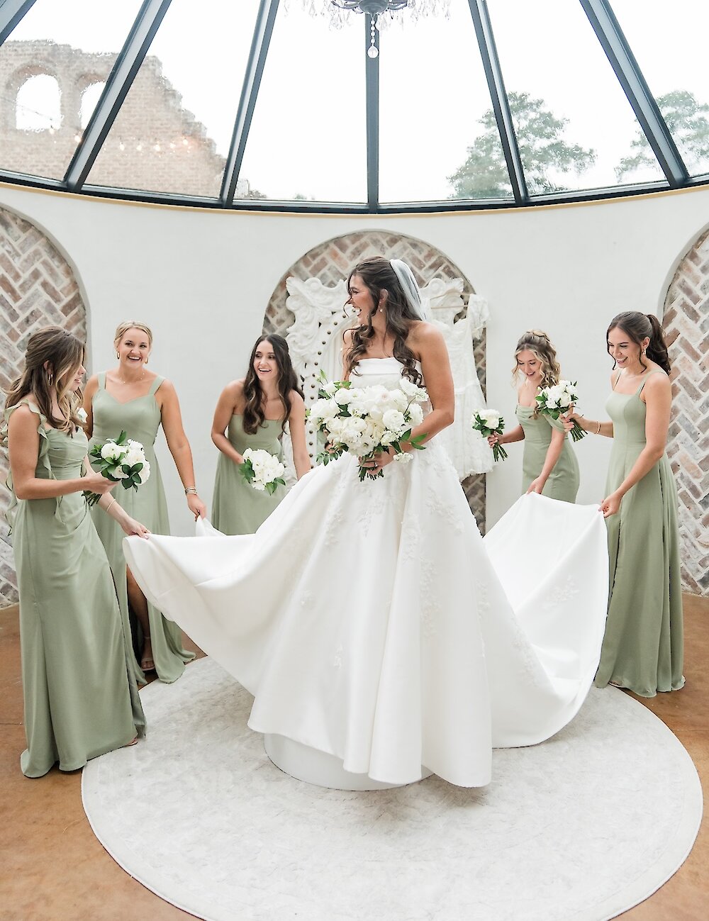 The smiling bride in her white ballgown is surrounded by five bridesmaids in sage green dresses, holding bouquets in a bright sunroom with a glass roof.