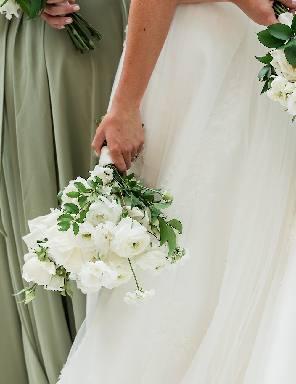 Close-up photo showing a bridal bouquet of white ranunculus and greenery held against the white bridal gown and a sage green bridesmaid dress.