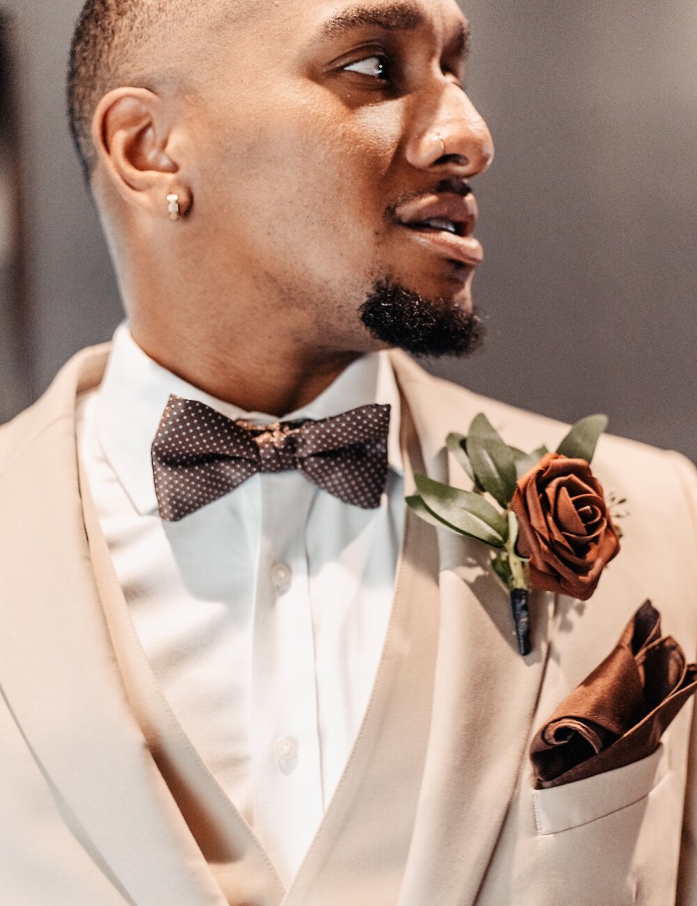 Close-up of the groom wearing a beige suit jacket, a polka dot brown bow tie, and a matching brown rose boutonniere.