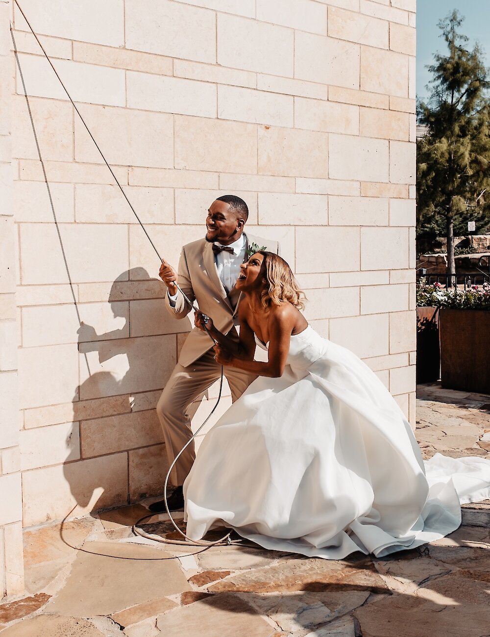 The bride and groom are laughing as they playfully pull on a rope to ring a bell against a light stone wall after their wedding ceremony.