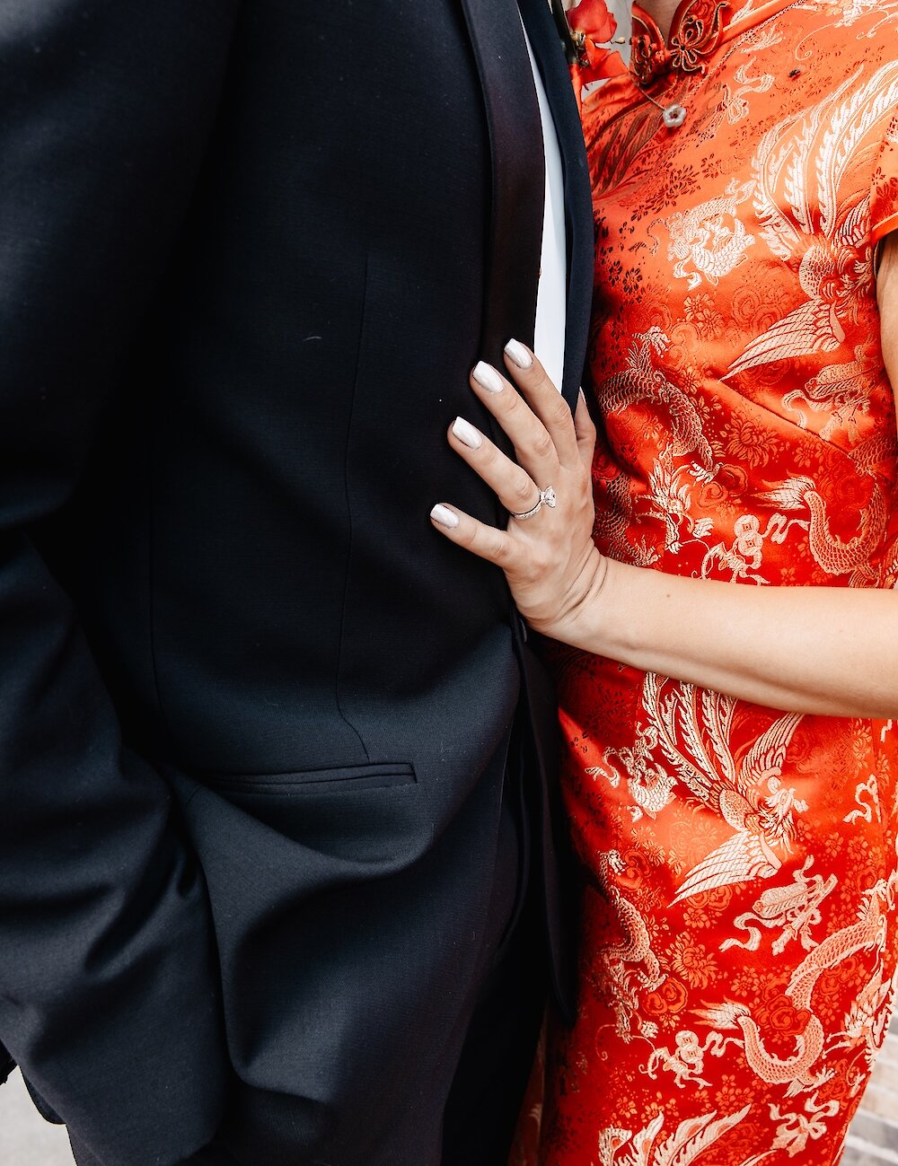 Close-up showing the bride in a vibrant red qipao with gold phoenix embroidery, with her hand holding the groom’s black tuxedo jacket.