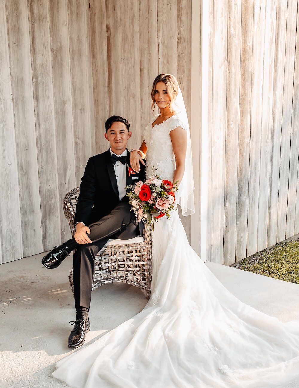 The groom sits in a wicker chair next to the standing bride in a lace cap-sleeve dress, holding a red and pink bouquet, against a rustic white wood wall.
