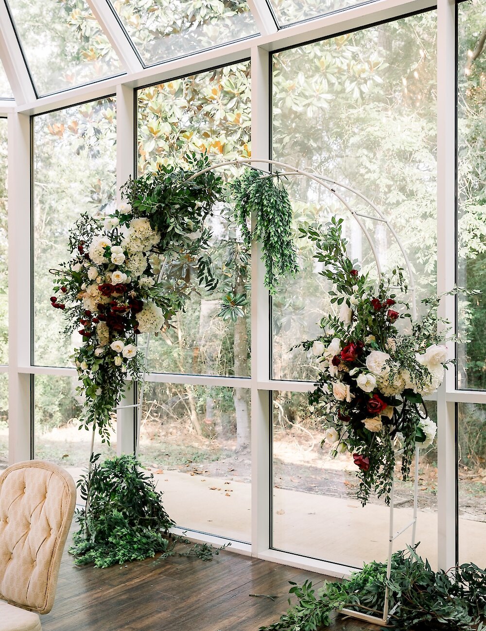 A white circular ceremony arch decorated with lush green and dark red florals stands in a bright room surrounded by tall glass windows.