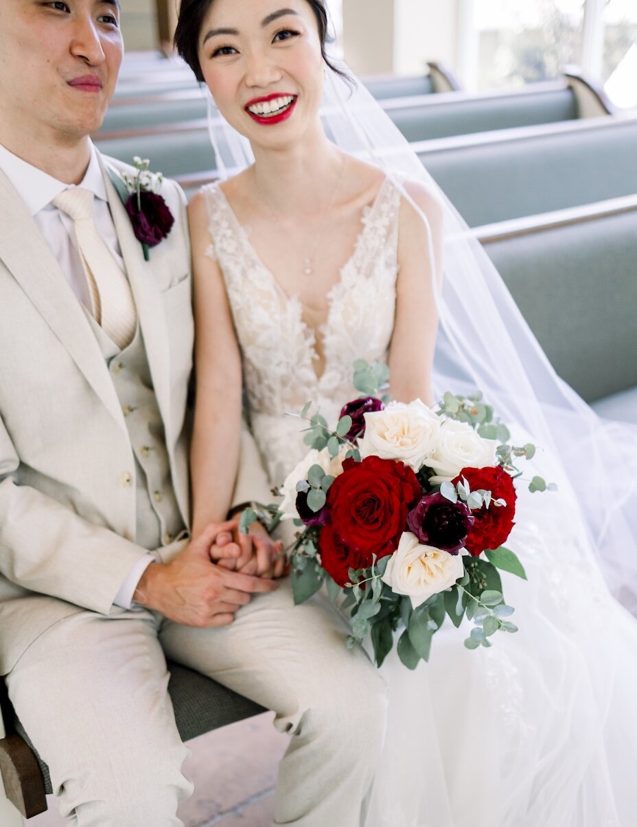 The bride and groom are sitting side-by-side, holding hands, with the bride holding a vibrant bouquet of red, white, and burgundy roses.