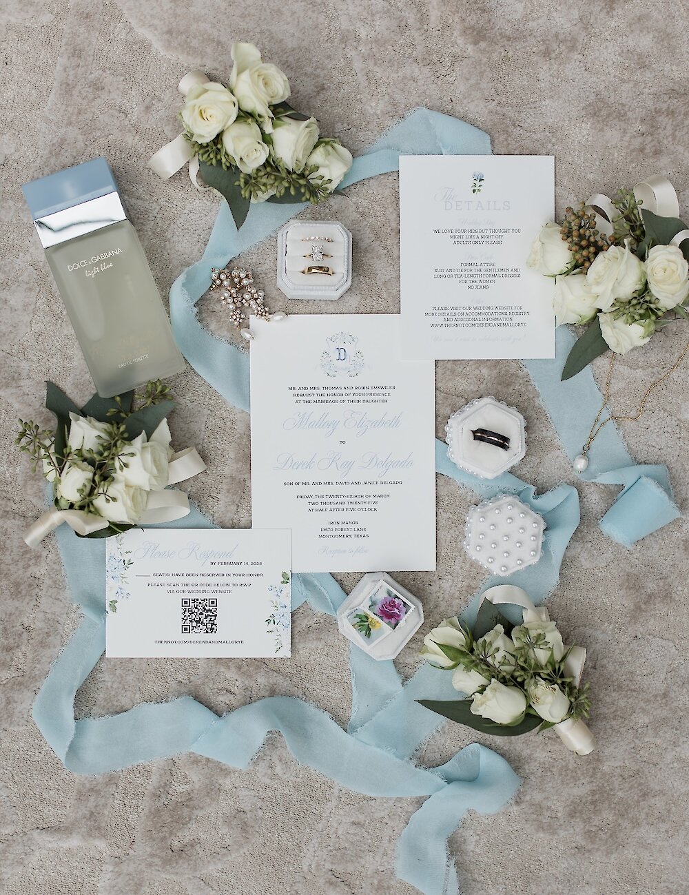 A wedding flat lay featuring the invitation suite, perfume bottle, rings, boutonnieres, and silk blue ribbon on a marble-style background.