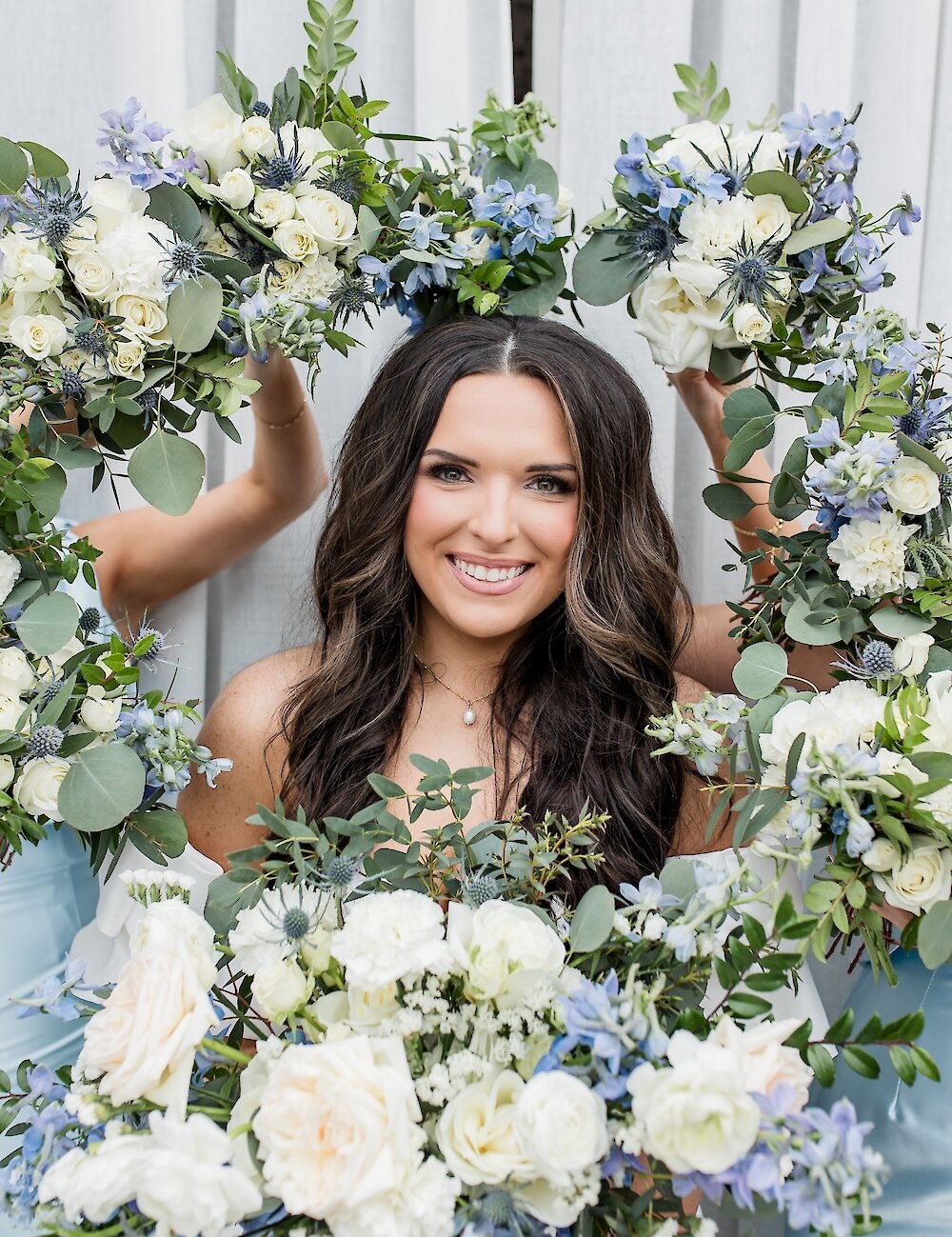 The smiling bride is framed by several bridesmaids' bouquets featuring white roses, blue thistle, and delphinium, set against a white draped background.