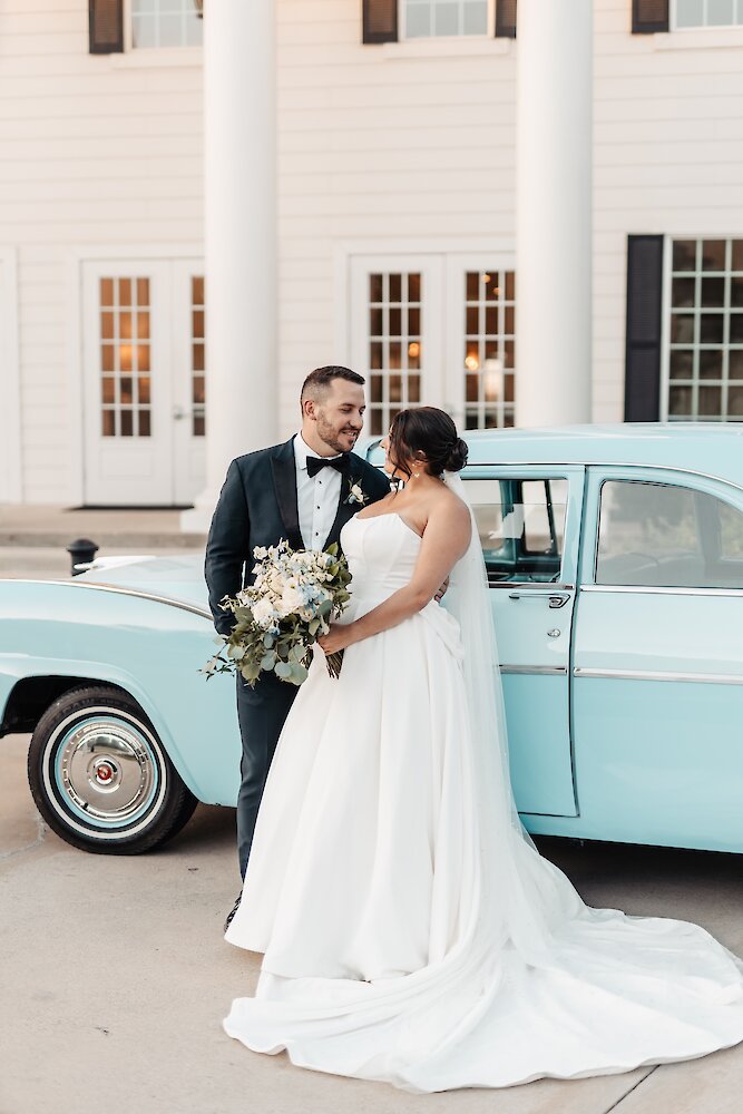 The bride and groom pose lovingly by a vintage light blue car in front of a white columned mansion venue.