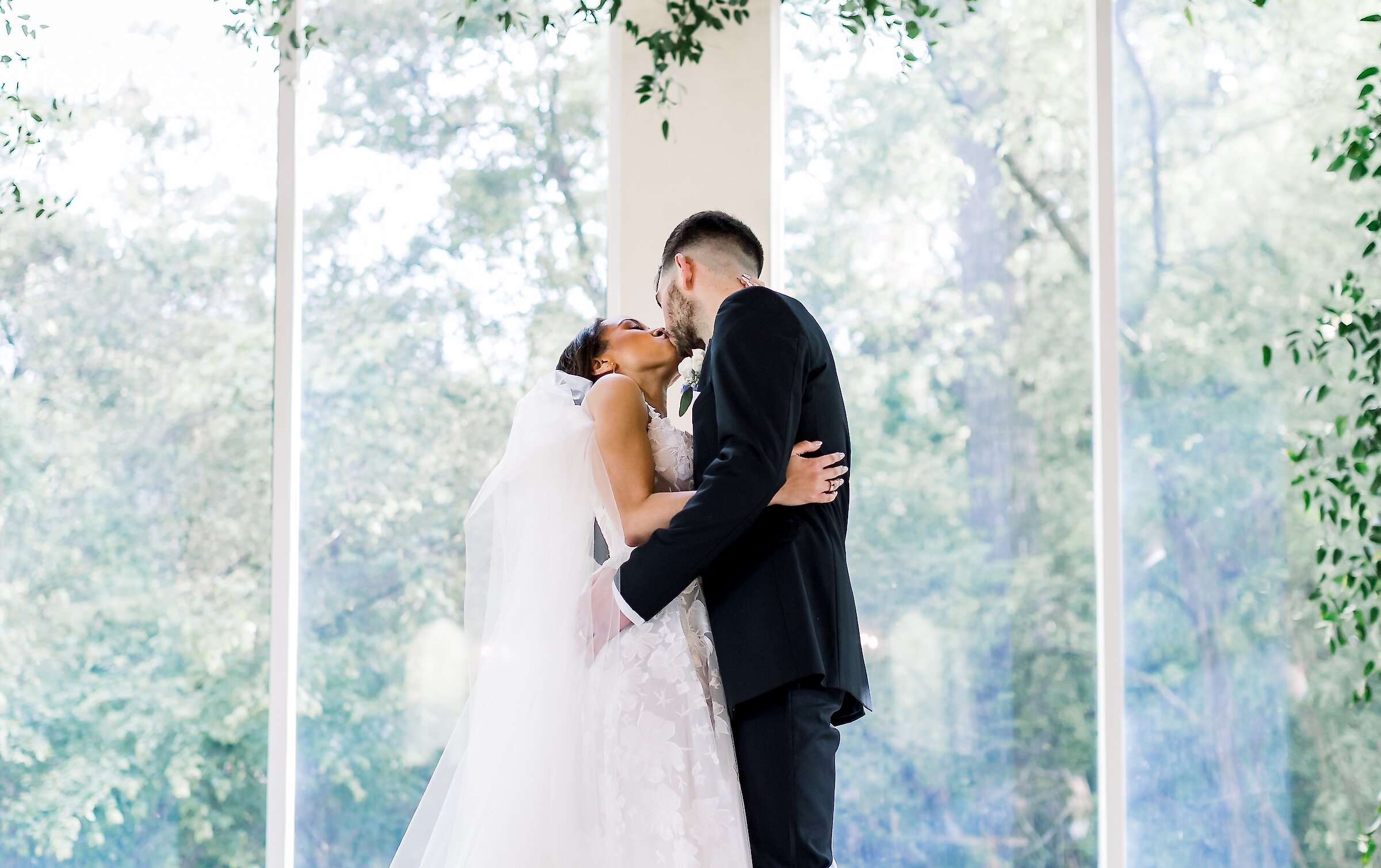 The bride and groom share a kiss under a suspended green floral arch, framed by tall glass windows overlooking a lush outdoor setting.