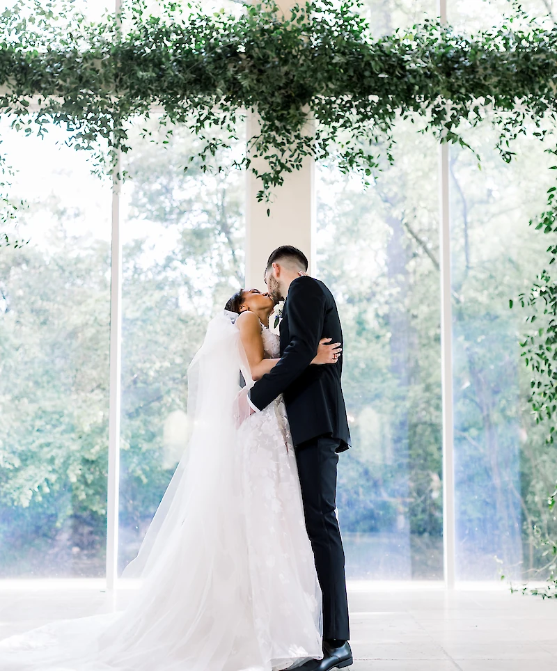 The bride and groom share a kiss under a suspended green floral arch, framed by tall glass windows overlooking a lush outdoor setting.
