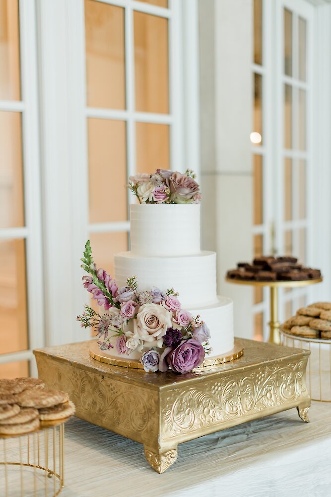A three-tiered white wedding cake decorated with cascading purple and mauve fresh flowers, displayed on a square gold stand.