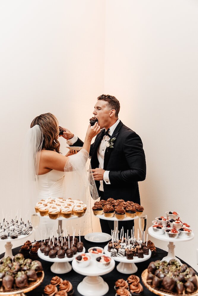 The bride and groom laugh while feeding each other chocolate cupcakes over a large wedding dessert bar with various sweets.