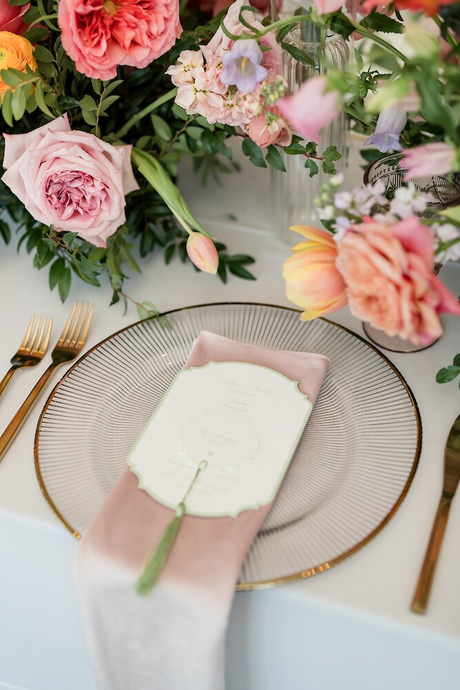 An elegant wedding place setting featuring a gold-rimmed plate, a pink velvet napkin, a decorative menu, and a vibrant floral centerpiece.