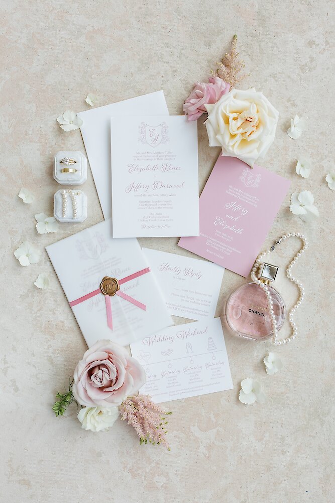 An elegant wedding invitation suite flatlay featuring pink calligraphy, rings, a pearl necklace, a wax seal, and light pink and white roses.