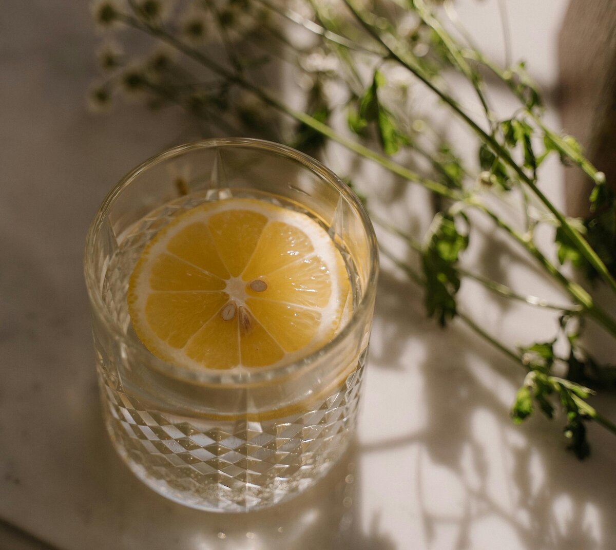 A close-up of a cut crystal glass filled with water and a bright yellow lemon slice, placed next to white daisy flowers in soft sunlight.
