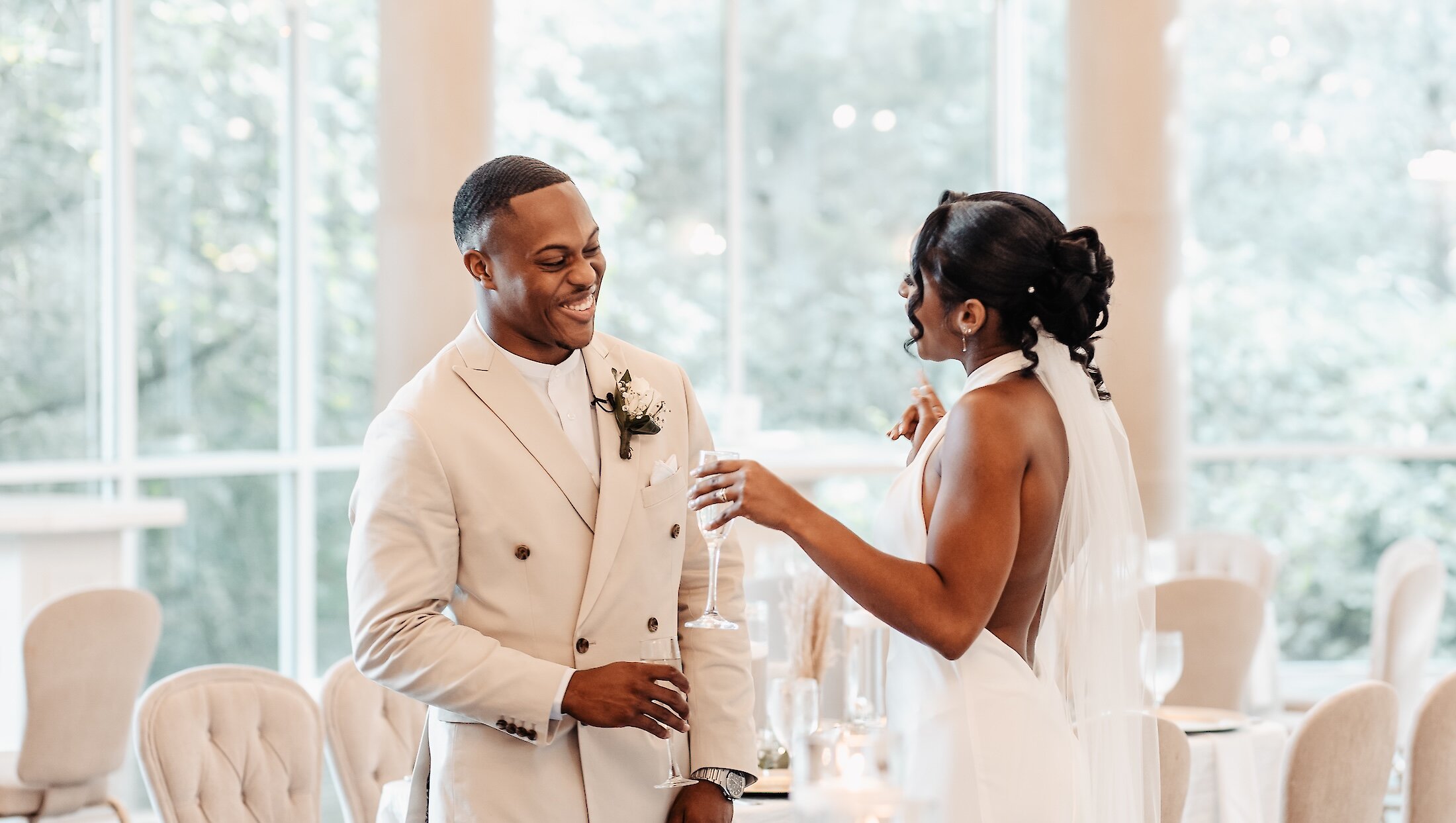 The smiling groom in a beige suit stands facing his bride, who is holding a champagne flute, inside a bright reception hall with large windows.