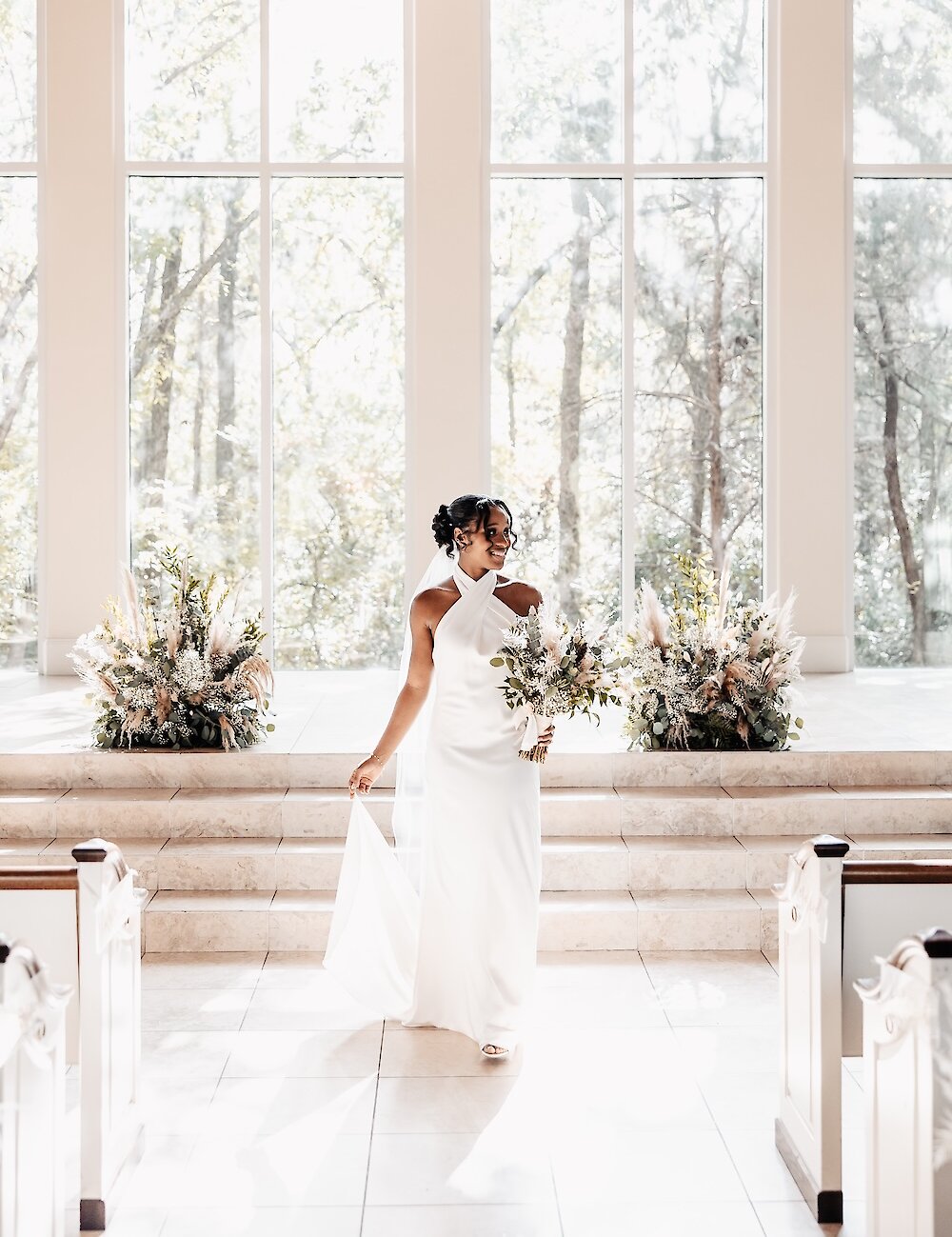 The radiant bride in a halter-neck white gown smiles while walking down the aisle of a chapel with tall windows and natural floral arrangements.