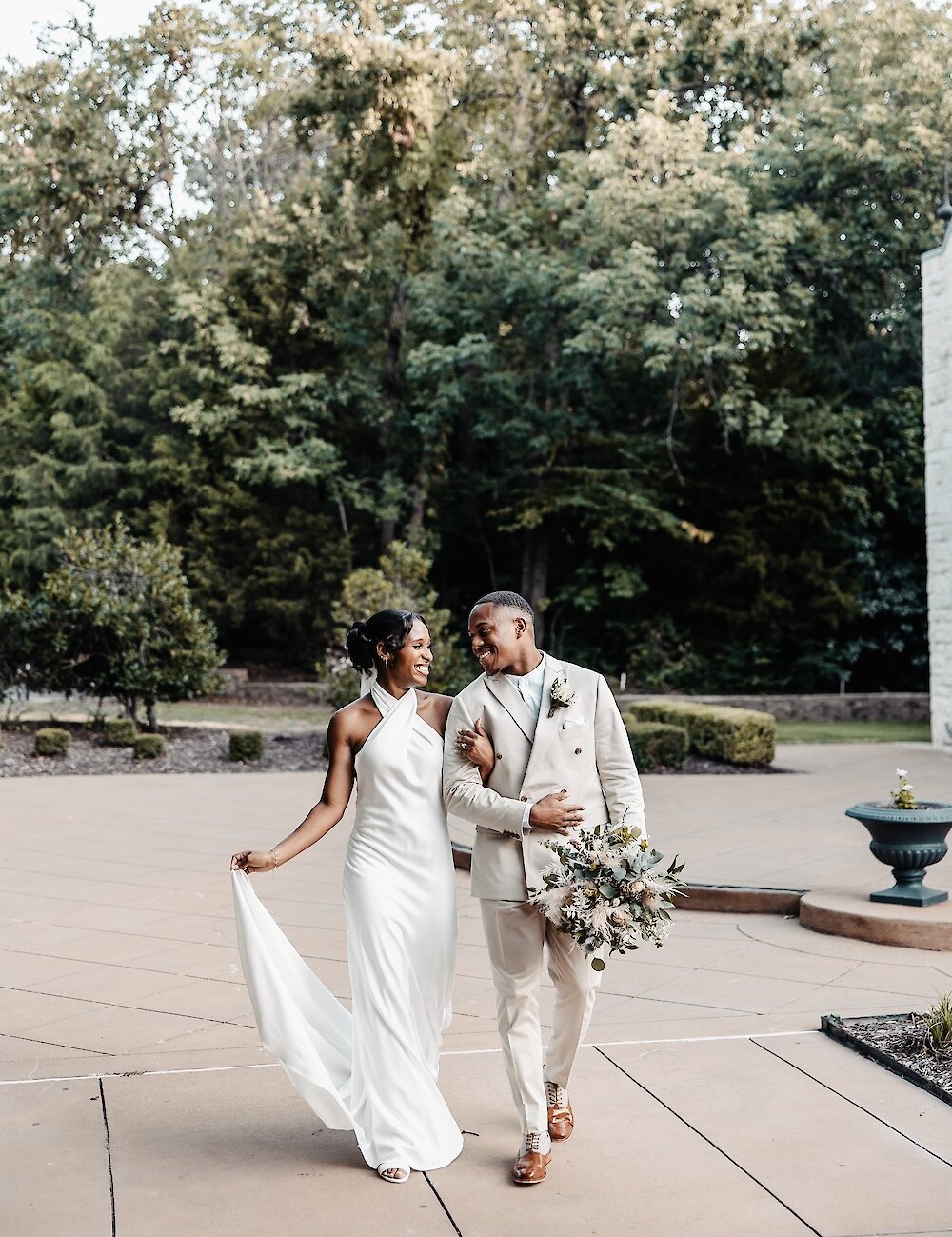 The newlyweds smile at each other while strolling outdoors, with the groom in a beige suit and the bride in a sleek white gown, holding a bouquet.