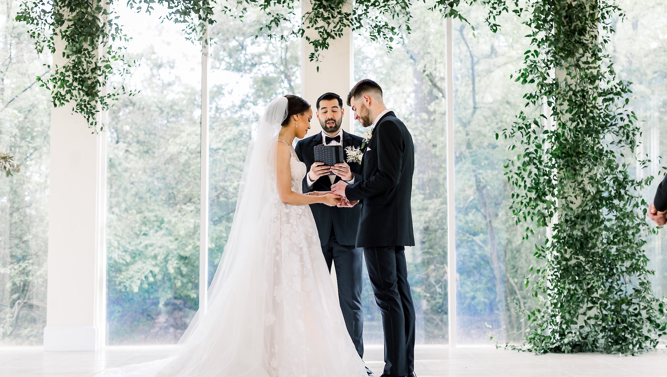 The bride and groom exchange rings with the officiant in a bright glass conservatory decorated with lush green vines hanging overhead and along the walls.