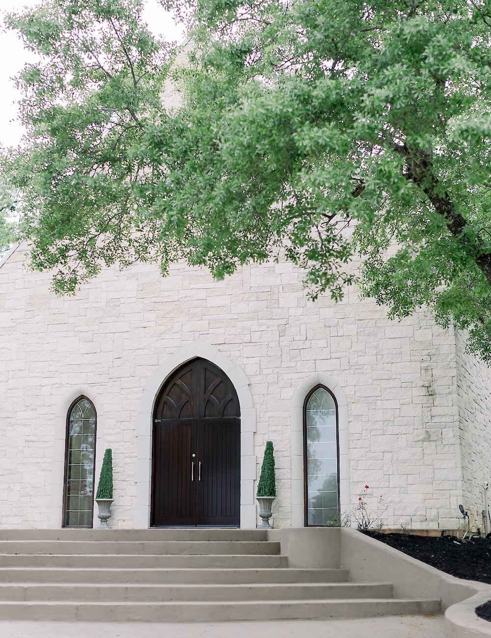 The exterior of a wedding chapel featuring a light stone facade, concrete steps, a dark gothic arched wooden door, and tall arched windows.