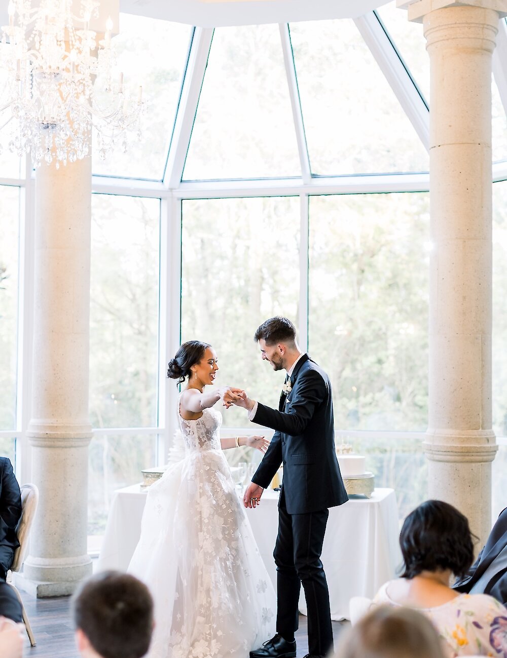 The bride and groom are happily dancing during their reception in a bright conservatory with stone pillars and a crystal chandelier hanging above them.