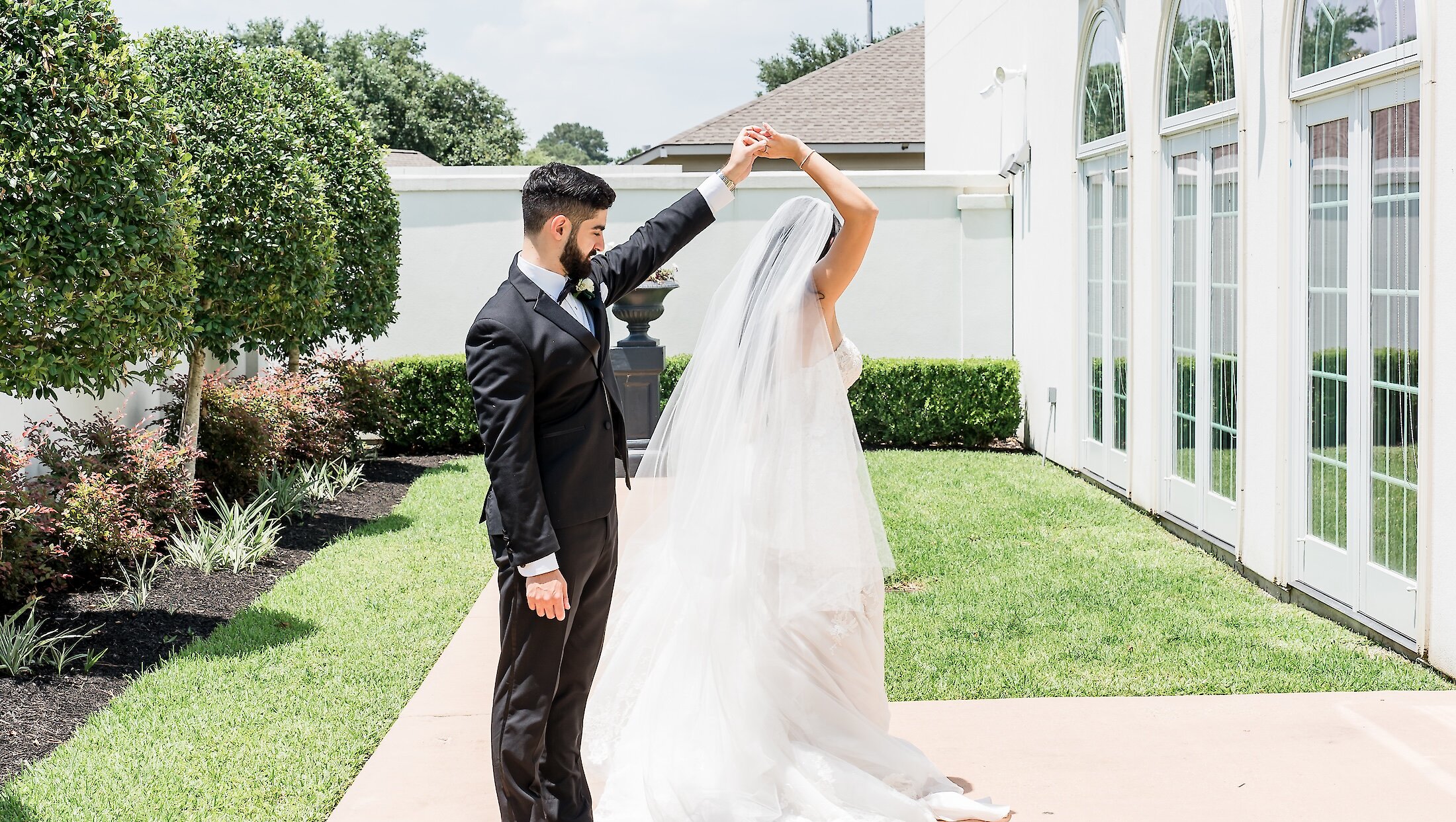 The groom twirls the bride on an outdoor patio next to a white building with arched windows and manicured green landscaping.