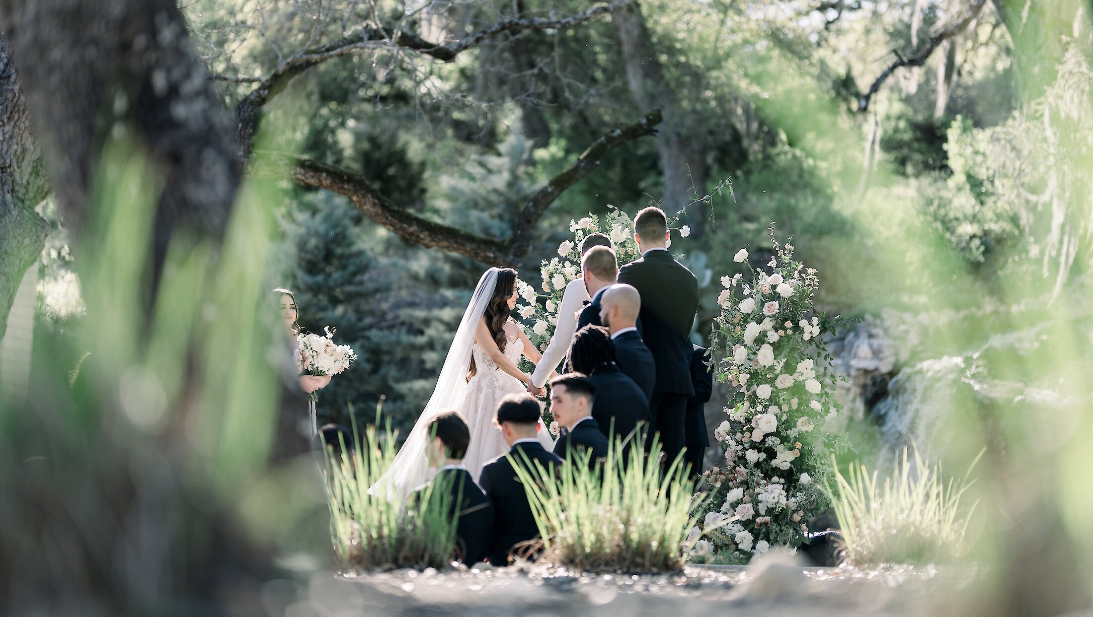 An intimate outdoor woodland wedding ceremony, viewed through foreground foliage, with the bride and groom at a floral arch in a clearing.