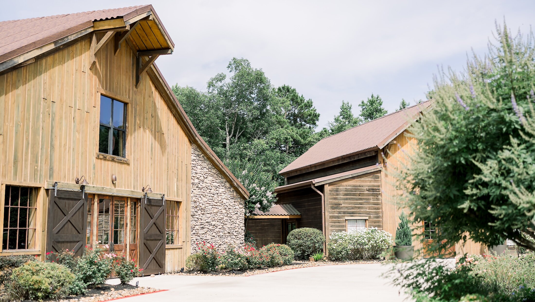 The exterior of a rustic wedding venue featuring light wood siding, stone accents, dark barn doors, and lush green landscaping.