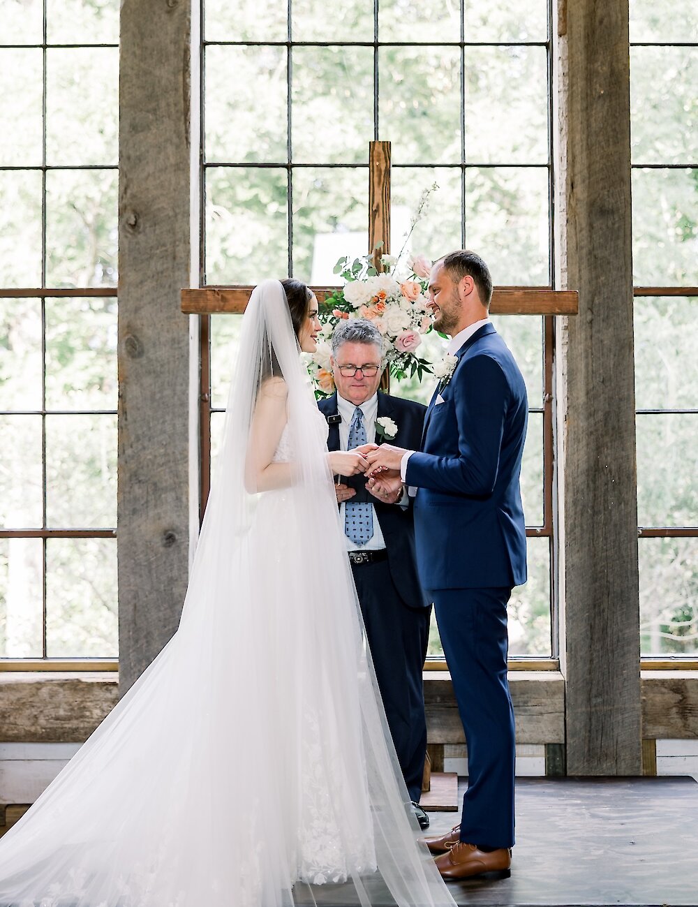 The bride and groom exchange rings at a rustic wooden cross altar, with the officiant presiding, in front of large paneled windows.