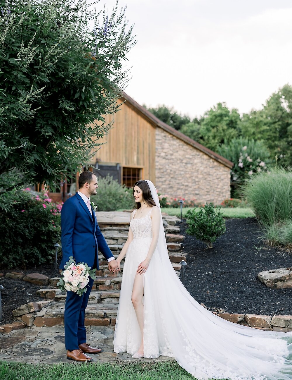 The bride and groom pose holding hands on a stone walkway outside their rustic barn venue, with the groom holding the white and pink bouquet.
