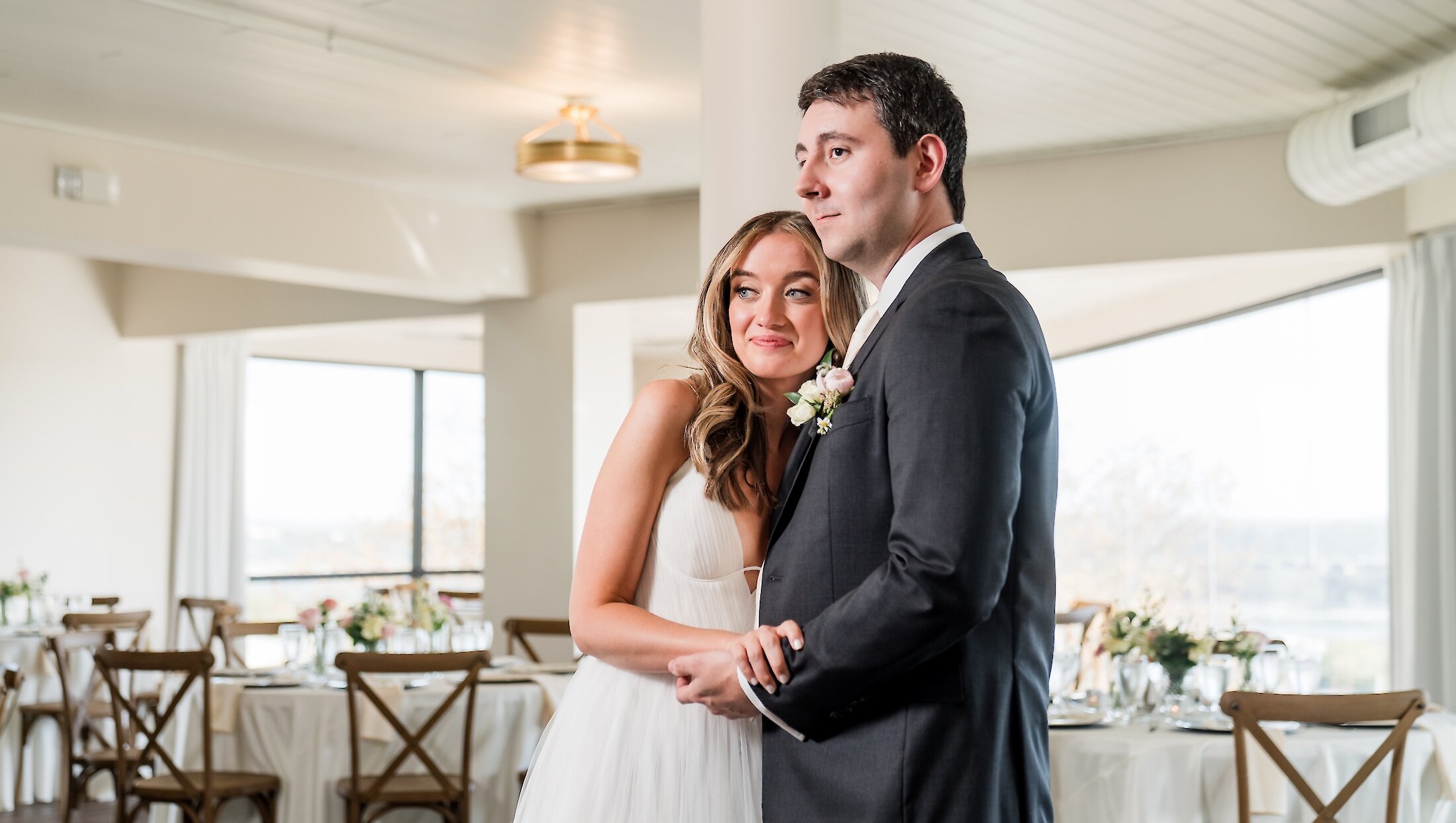 The bride hugs the groom while posing for a romantic portrait in their bright, modern reception hall with cross-back chairs and large windows.