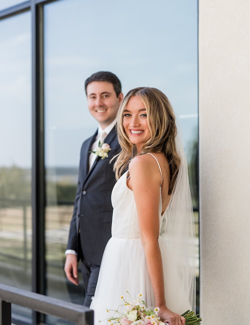 The smiling bride, holding a multi-colored wildflower bouquet, looks over her shoulder while posing with her groom on a sunlit balcony.