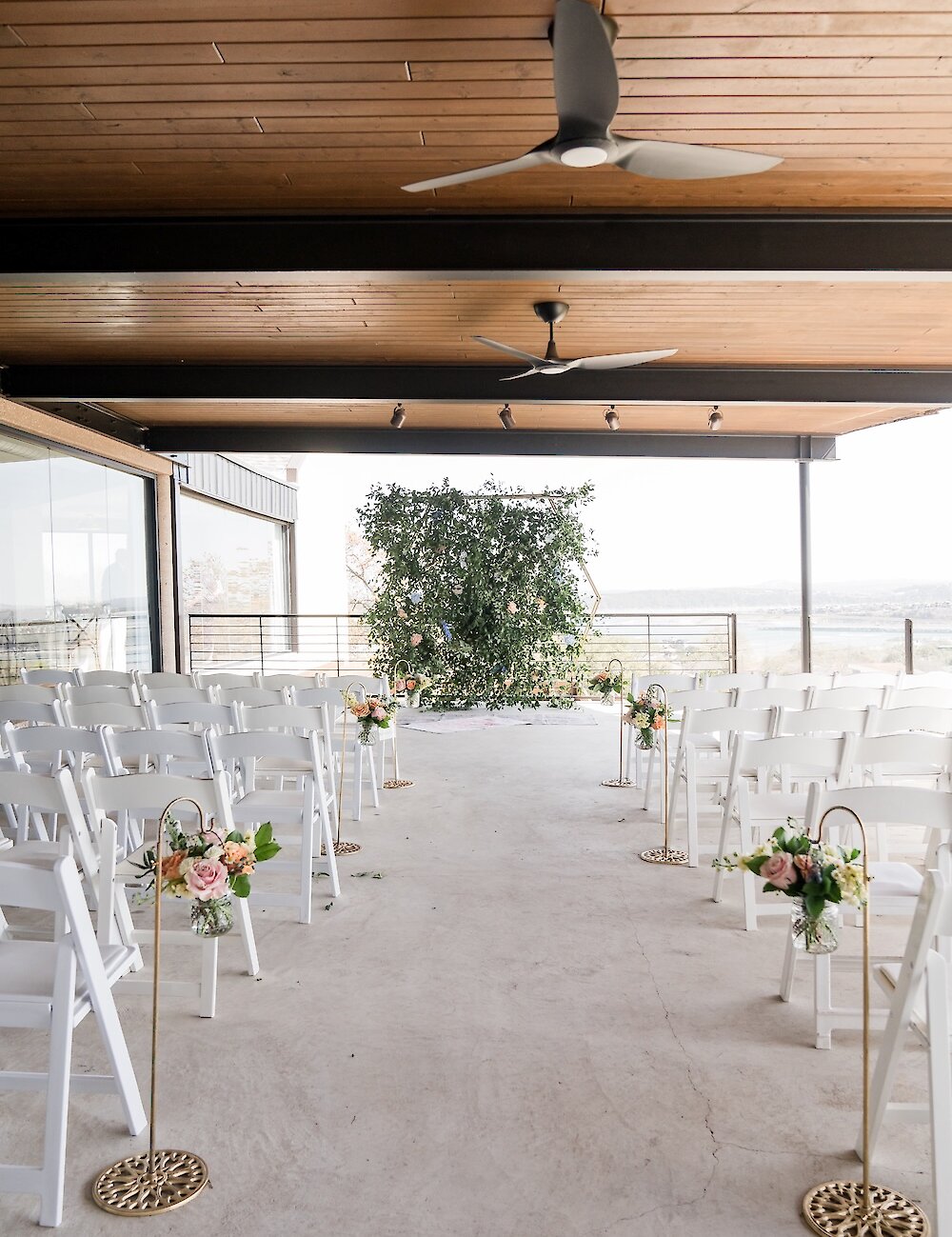 An outdoor covered ceremony patio with white folding chairs, a green backdrop, and a distant view of the landscape and water.