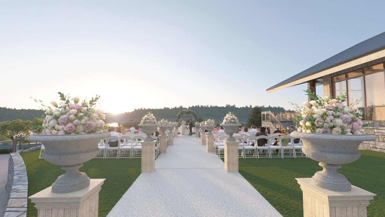 An outdoor wedding ceremony setup on a grassy lawn at sunset, featuring a white aisle, white guest chairs, and large stone urns filled with pink and white flowers.