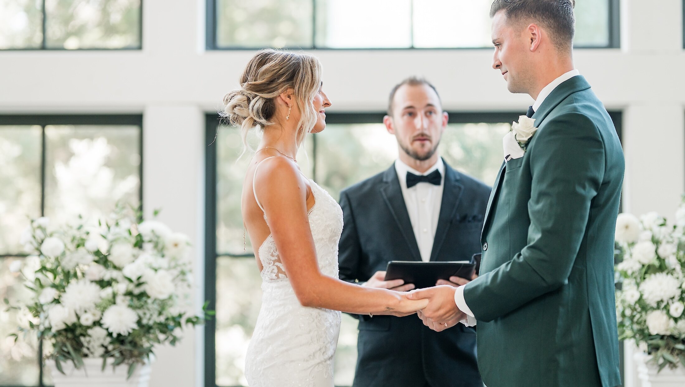 The bride and groom hold hands and look at each other while exchanging vows with the officiant in a bright ceremony room with large windows.