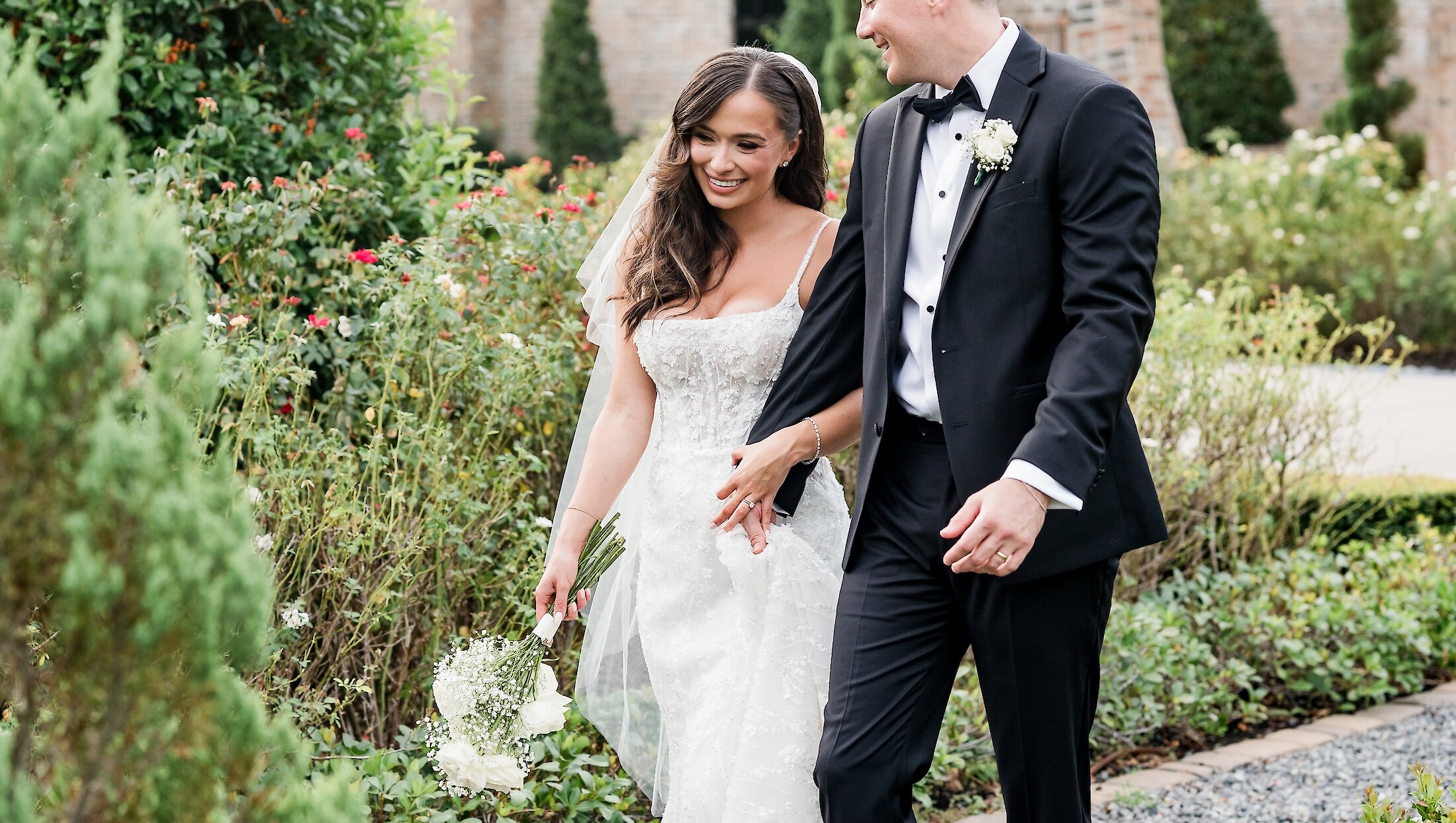 The smiling bride, holding a white bouquet, walks arm-in-arm with her groom through a lush green and flower-filled garden path.