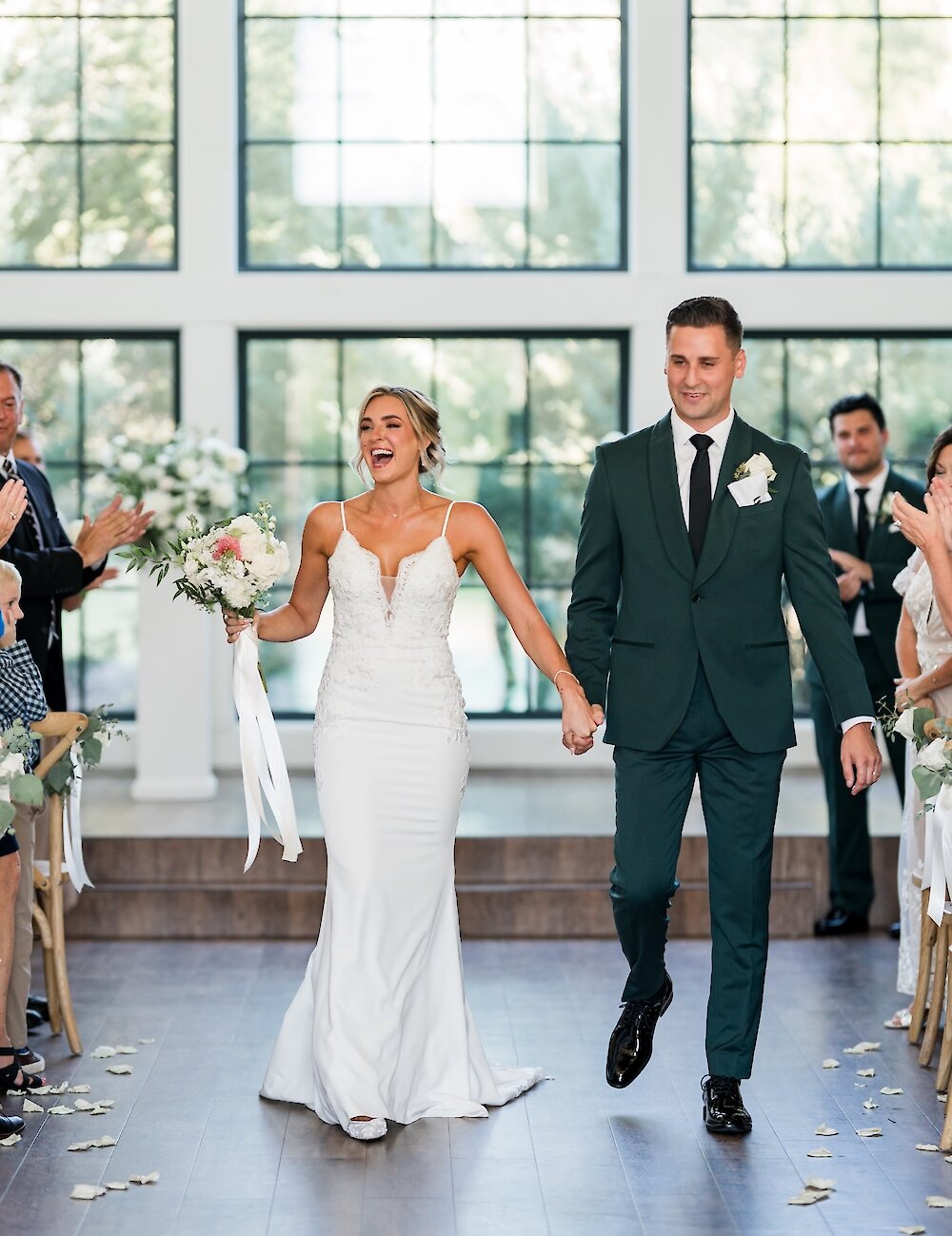 The ecstatic newlyweds hold hands and smile while walking down the dark wood aisle after the ceremony, with the groom wearing a dark green suit.