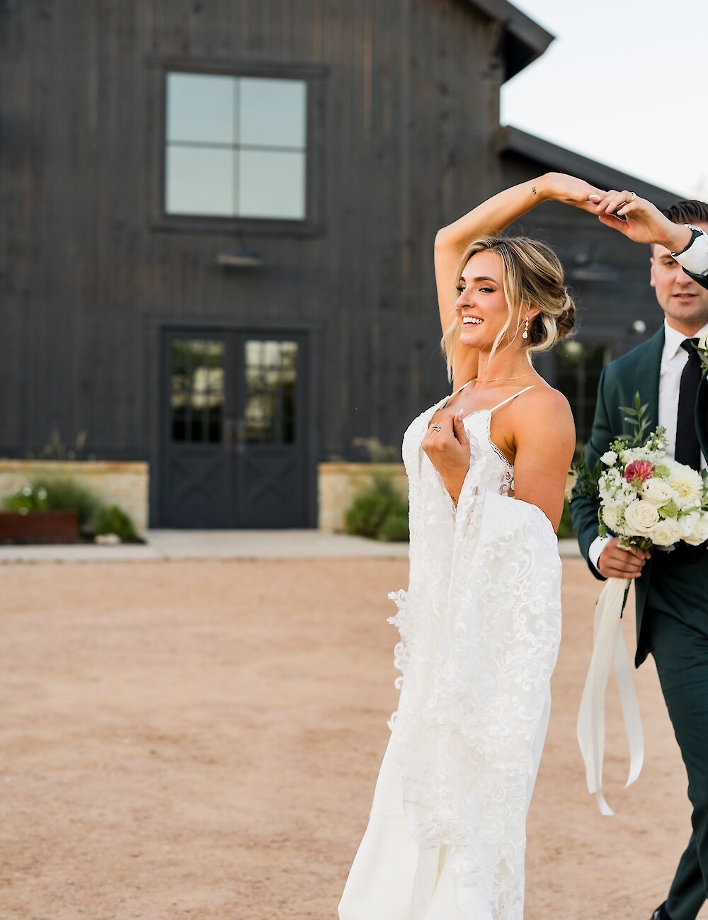 The joyful bride is twirled by her groom in front of a modern black barn venue, wearing a lace gown and holding her dress.