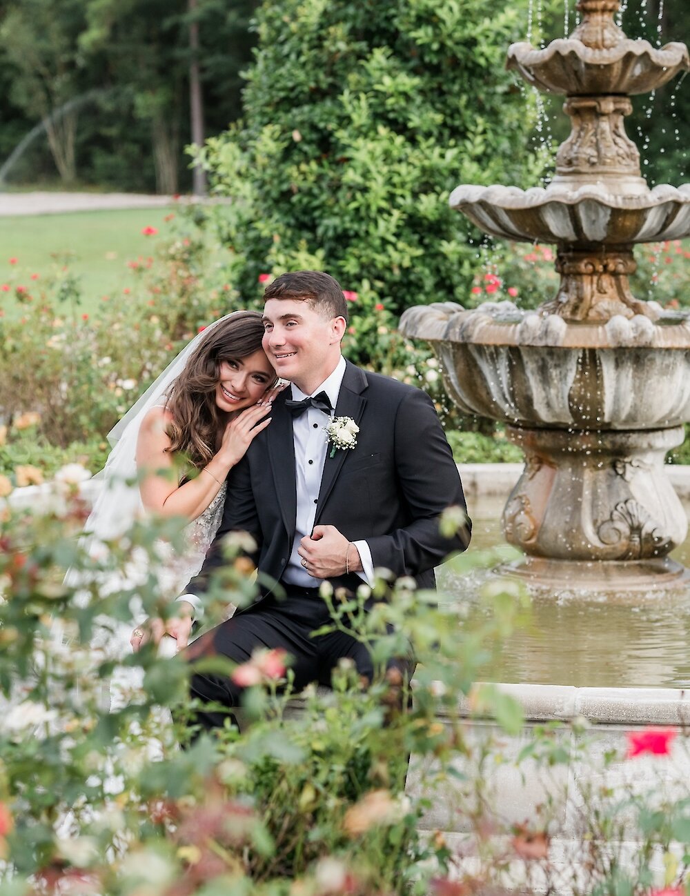 The bride sweetly rests her head on the groom's shoulder while sitting near a tiered fountain in a rose garden during their wedding portrait session.