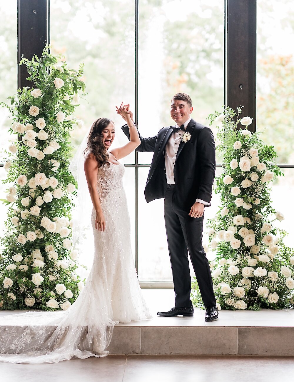 The excited bride and groom raise their hands in celebration, standing between two tall arrangements of white flowers in front of a large window.