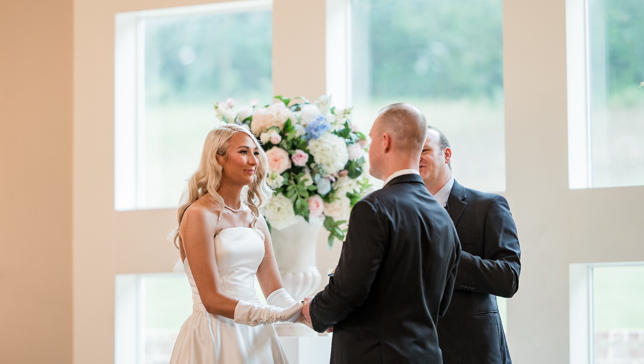 The blonde bride in a strapless dress and white gloves holds hands with the groom during their ceremony, centered around a large floral vase.
