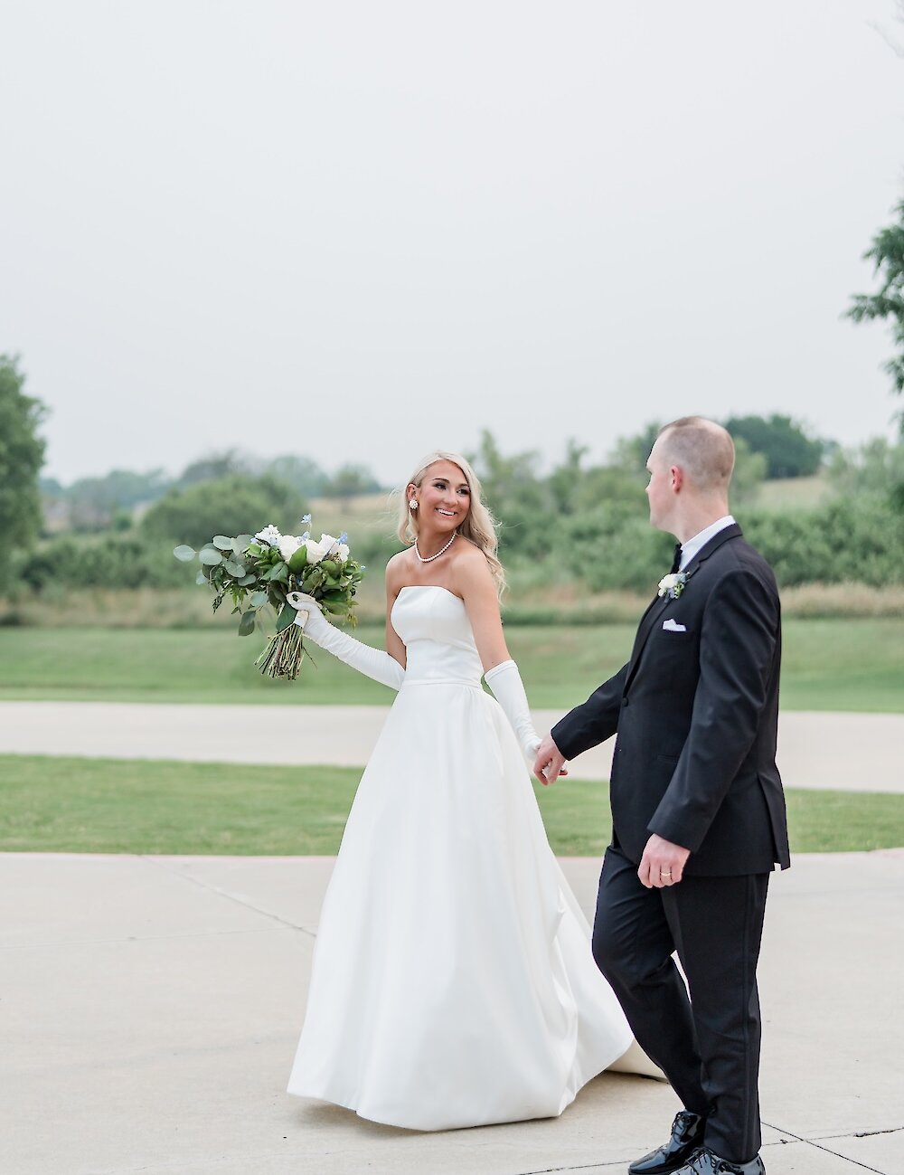 The smiling bride in a strapless gown and white gloves holds her bouquet while walking hand-in-hand with her groom on a concrete path outdoors.