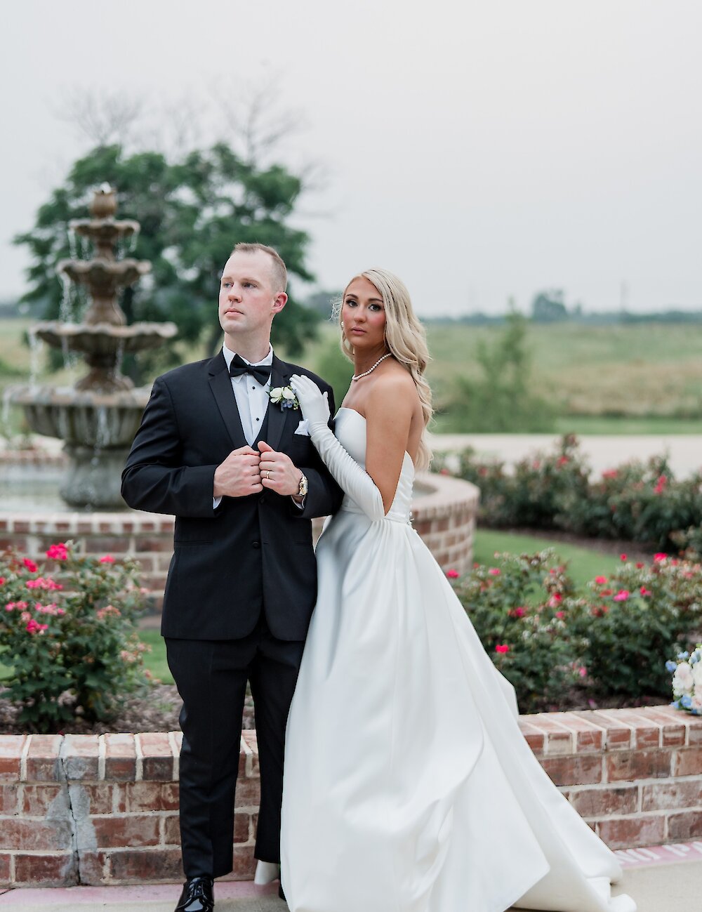 The bride in a satin gown and long gloves poses with her groom in a tuxedo next to a brick wall with a tiered fountain in the background.